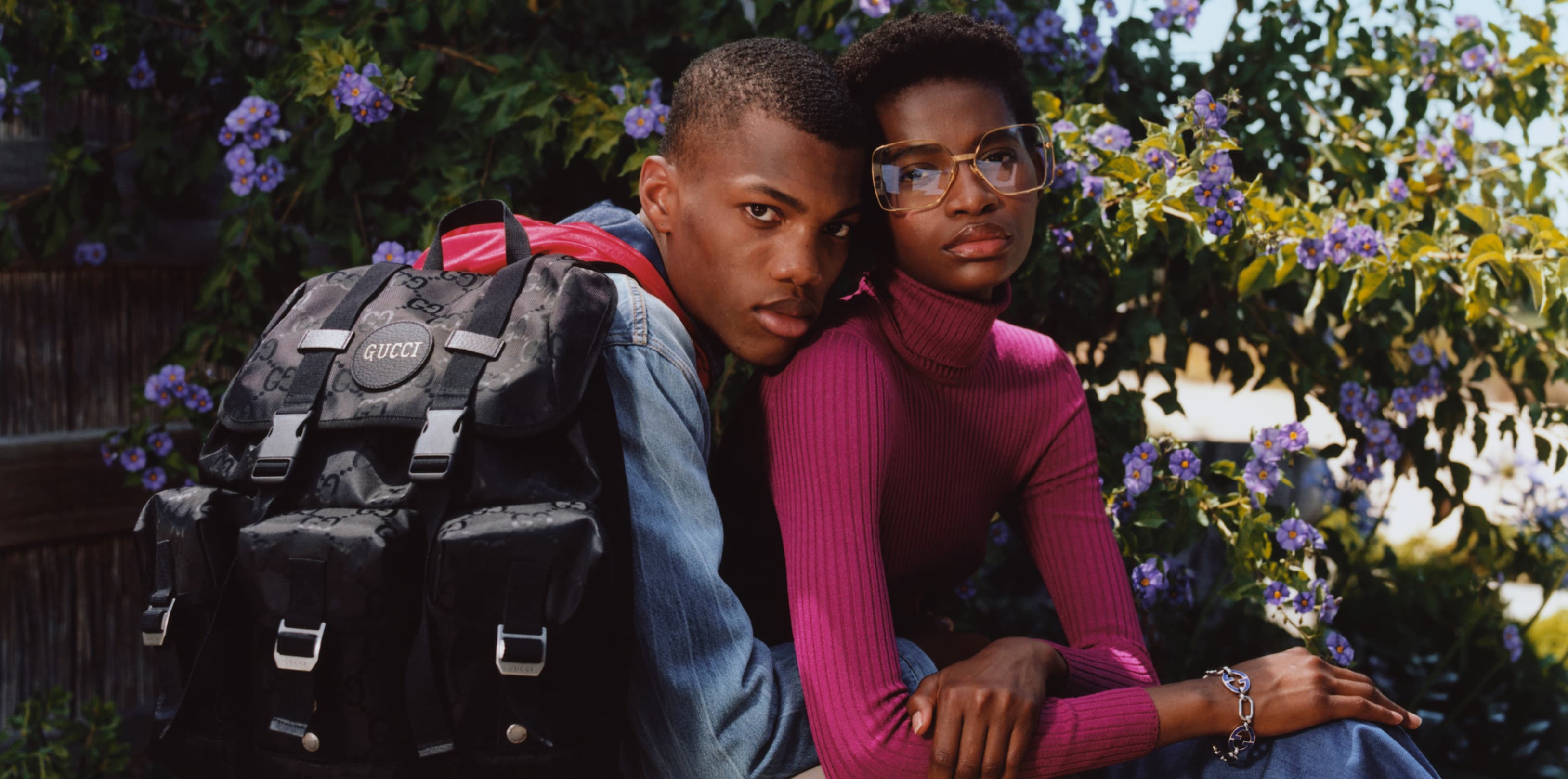A young Black man wearing a black regenerated nylon GG pattern backpack is seated in a garden with his arm around the waist of a young Black woman. Behind them is a large Paraguyan nightshade bush with purple and yellow flowers as well as a weathered wooden fence. He is wearing a denim jacket with beige pants, and she is wearing a raspberry colored ribbed turtleneck and oversize square metal frame eyeglasses.