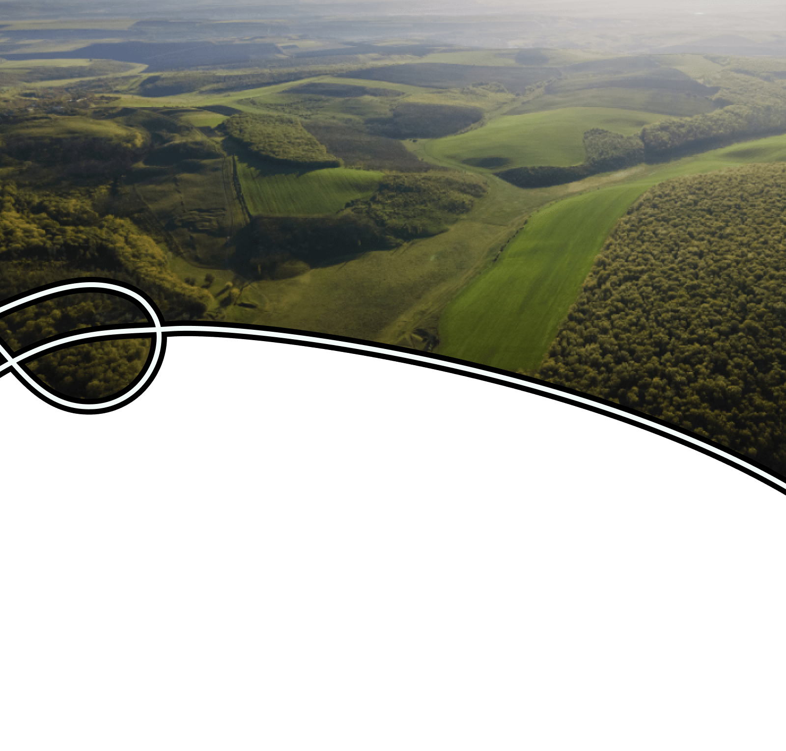 In an aerial image, a slight haze clings to a lush green landscape with rolling hills under a blue sky. The landscape is a mix of dark green forest and irregular bright green patches carved out by agricultural activity.