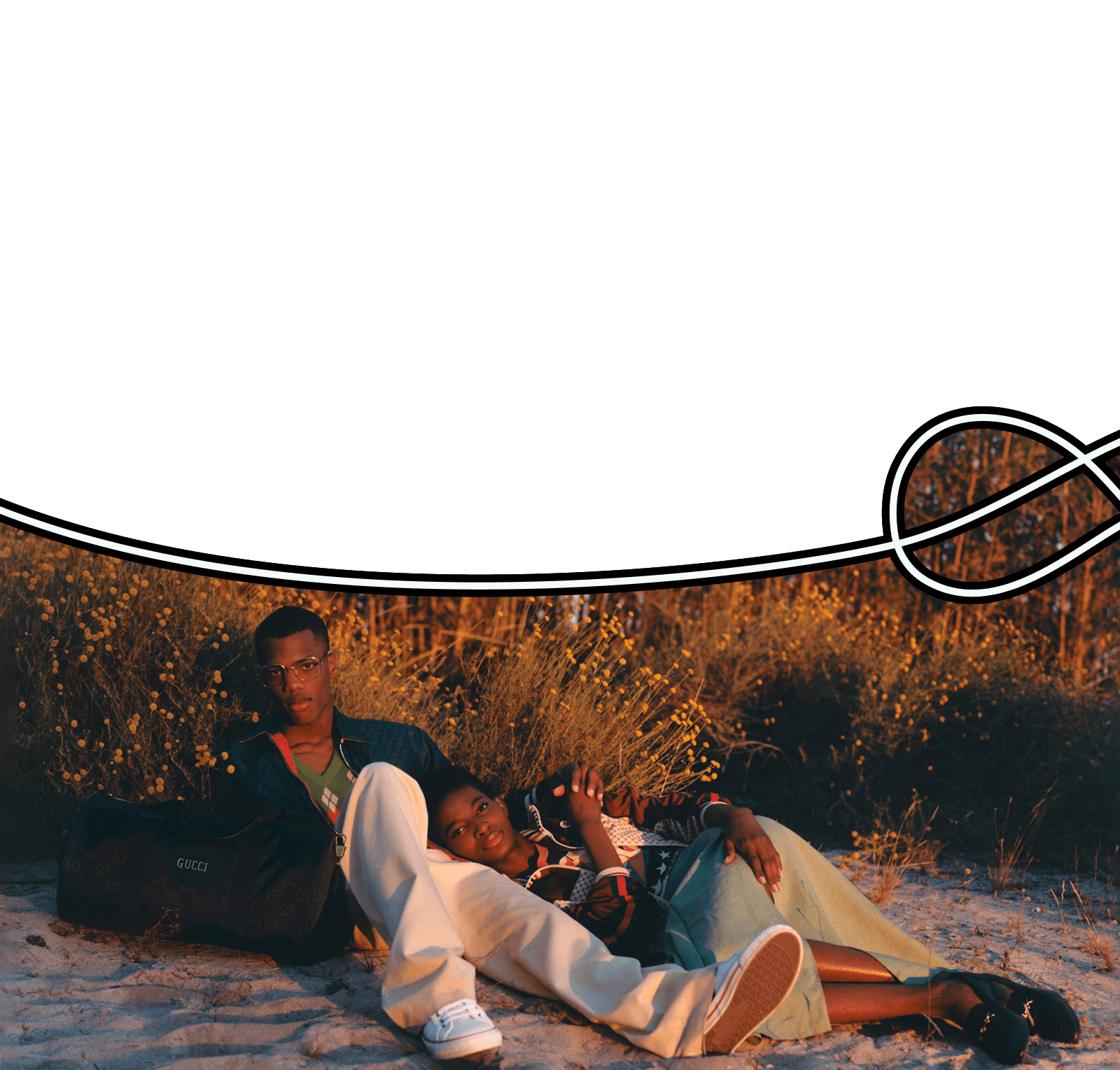 Next to a large regenerated nylon black GG duffle bag, a young Black man reclines on a sandy dune with his arm around the shoulder of a young Black woman, lying with her head resting on his hip. The top of the frame is filled with dense pine trees in the distance and there are shrubs with small yellow flowers directly behind them. He is wearing a navy track jacket over a green argyle sweater, with ivory pants and white sneakers. She is wearing a multicolor track jacket and sweater with navy loafers and a sea foam green skirt.