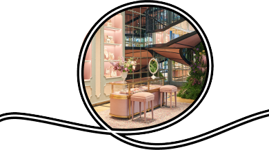 The interior of a high ceiling Gucci store features a very large spiral staircase surrounded on two sides by with mirror paneled walls, a parquet floor, and pale gray and pale pink display wall with decorative trim and well lit shelves full of white handbags a nd other small accessories. A large column of plants, including wisteria, grows up the middle of the staircase, and there is a small display case island on a Persian rug with small pink stools and a white vanity mirror. On the left side of the frame are tw o mannequins in long dresses, one floral, one pale pink, under an archway that leads into another part of the store.
