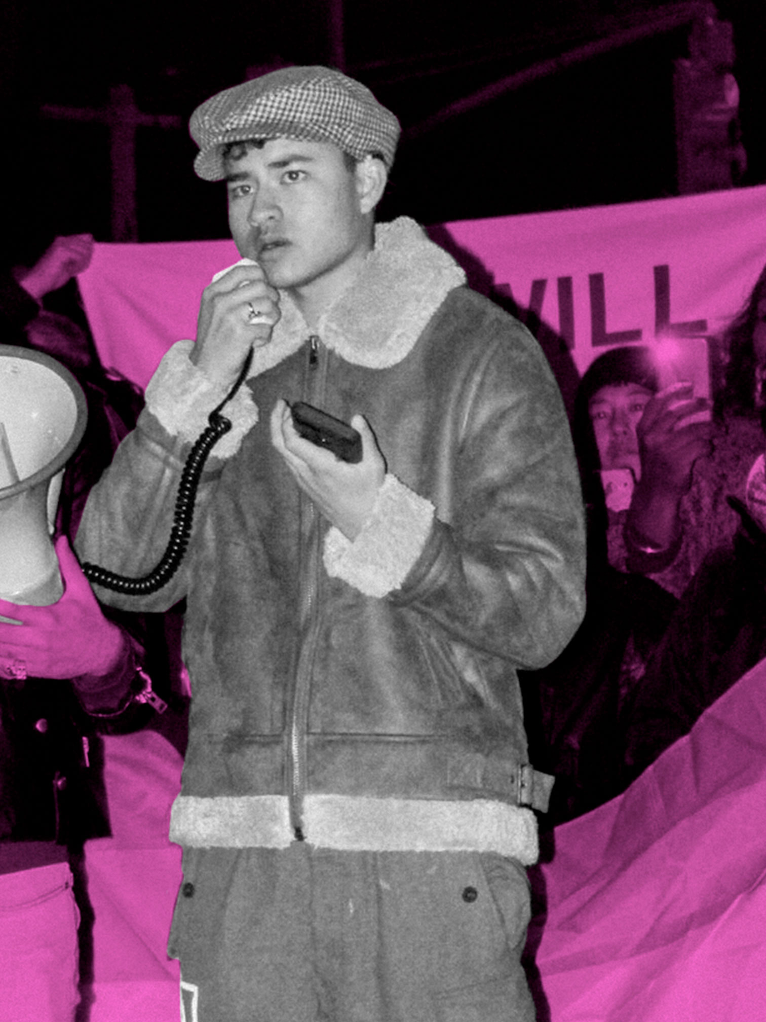 Chella Man speaks on stage at a protest. He wears a moto jacket, hat, and pants, and holds the mic to a megaphone, looking out at the crowd. The image is filtered in neon pink, while Chella Man remains filtered in black and white.
