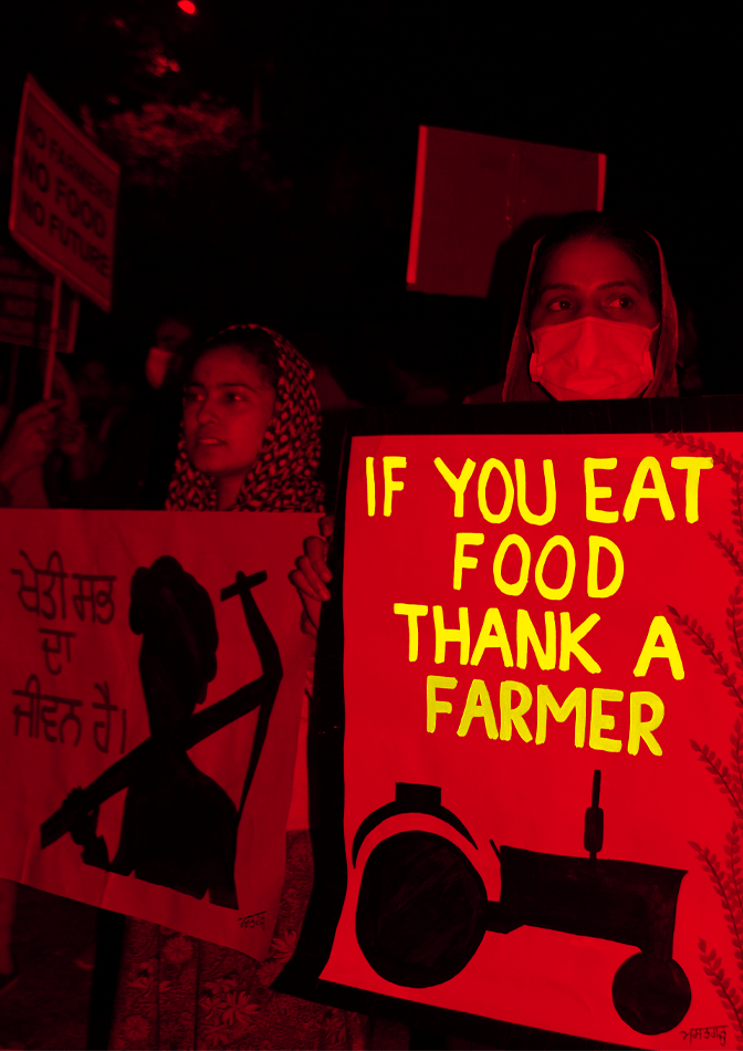 Two women hold signs. The first woman is wearing a headscarf and holding a sign written in Hindi. The second woman is wearing a headscarf and a mask. She is holding a sign with a drawing of a black tractor and the words “IF YOU EAT FOOD THANK A FARMER.” The sign and text alternate between red and yellow, while the background and women are filtered in red.
