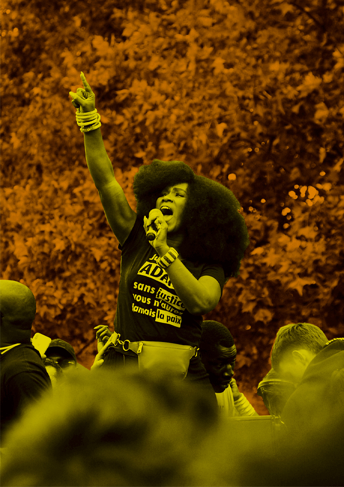 Assa Traoré stands amongst a crowd while shouting into a microphone and pointing to the sky with one finger. She wears bangles on both of her wrists, a belt bag, and a black t-shirt with white lettering that says “Justice and Truth for Adama, without justice you will never have peace” in French. The background and her silhouette alternate between yellow and orange.