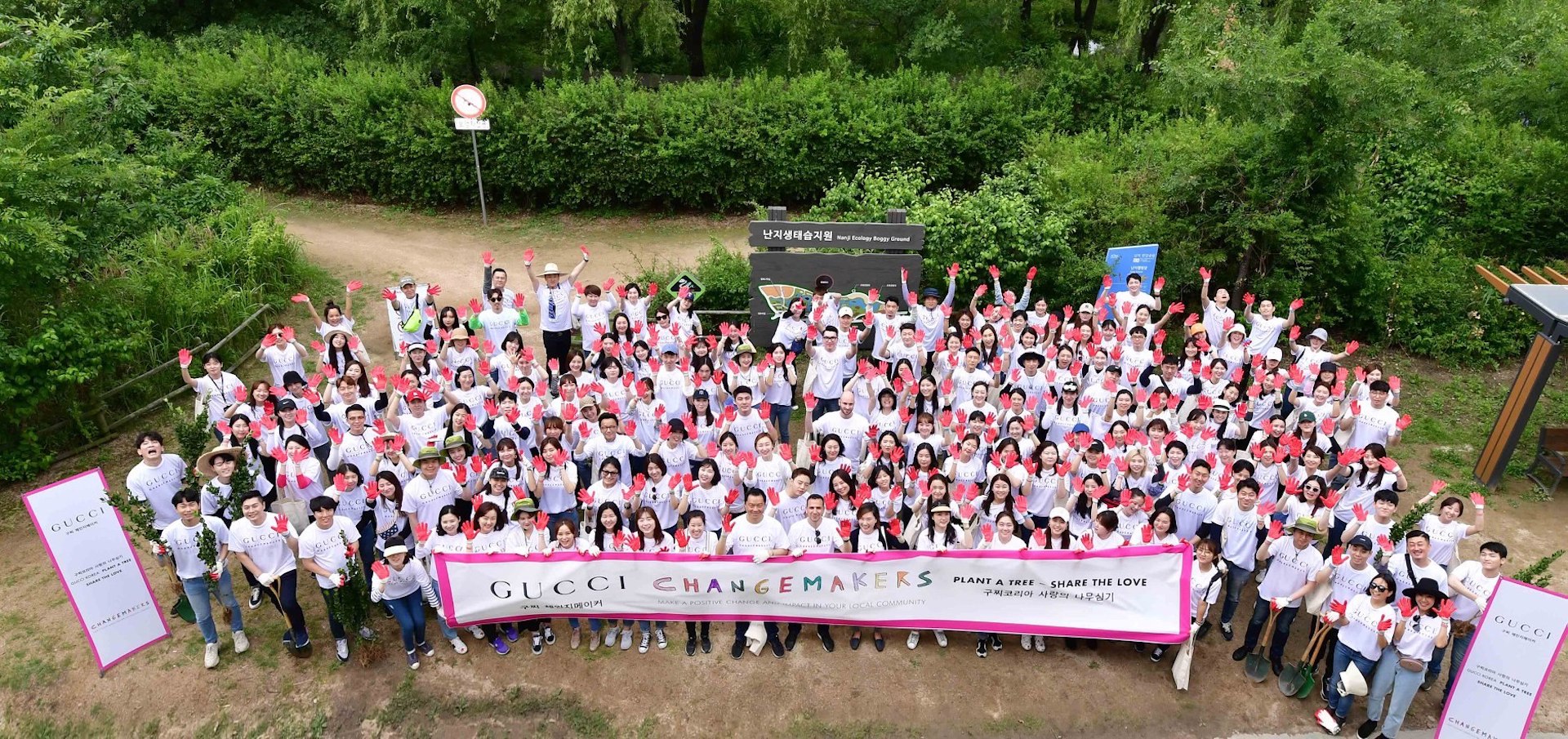 A large group of people wearing white shirts and red gloves photographed from above, look into and wave at the camera, holding a sign that says “Gucci Changemakers, plant a tree, share the love”. They stand on a dirt path in front of leafy green bushes and trees.
