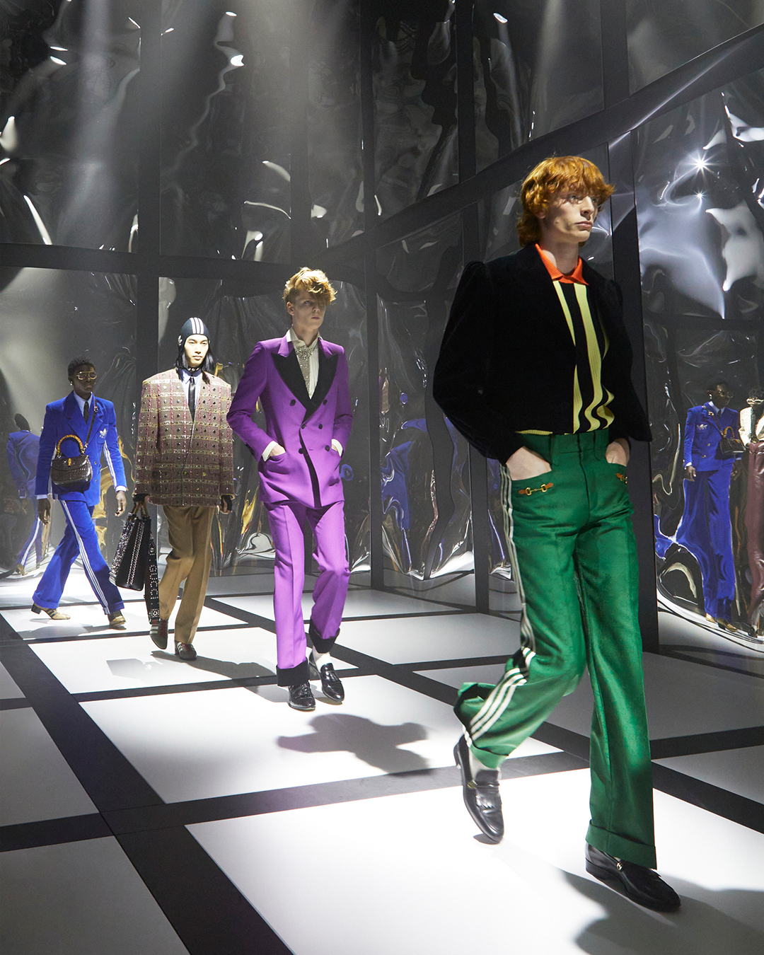 A row of four models walking the black and white grid runway of the Exquisite Gucci fashion show wearing various looks from the new collection. There are funhouse-style mirrors along the walls.